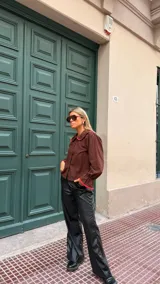 Camisa de color marrón chocolate con cuello Peter Pan y puños con volados, ambos detalles resaltados con un ribete en color rojo. Presenta cierre frontal con botones rojos.