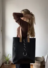 Tote bag de gran tamaño confeccionada en ecocuero negro con textura de reptil. Presenta una tira central decorativa con ojales metálicos y una argolla grande en la parte inferior, además de manijas en color gris.