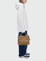 Bolso tote Marc Jacobs modelo The Jacquard, confeccionado en tejido de lona negro con detalles en blanco. Presenta el logo de la marca y la inscripción 'THE TOTE BAG' en el frente, con doble asa superior y correa ajustable extraíble.