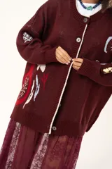 Saco de punto color vino con diseño holgado, cuello redondo y mangas largas. Presenta un gran bordado central en la espalda con forma de flor en tonos crema y rojo, acompañado de un pequeño bordado de sol y una frase bordada en la parte inferior.