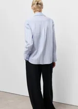 Camisa de corte recto con diseño de rayas verticales en tonos celestes y blancos. Presenta cuello camisero con solapas amplias, cierre frontal de botones ocultos bajo tapeta y mangas largas con puños abotonados.