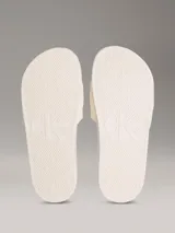 Ojotas tipo slide de color blanco hueso, con tira ancha acolchada y monograma de CK en relieve. La suela es de espuma.