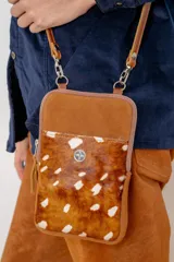 Bolso tipo pocket bag confeccionado en cuero vacuno con frente de pelo natural estampado con manchas blancas. Cuenta con cierre superior, bolsillo frontal con detalle de bordado metálico de flor de lis y correa de cuero desmontable.