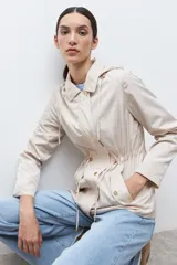 Campera impermeable color beige claro, con capucha desmontable, cierre frontal, cordón ajustable en la cintura y botones dorados.