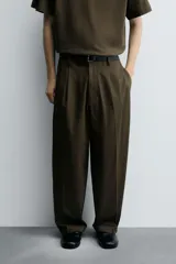 Pantalón negro de corte relaxed fit y estilo wide leg, con pliegues frontales dobles, bolsillos delanteros y detalle de bolsillos traseros de vivo. Cierre frontal cruzado con cremallera y gancho metálico oculto.