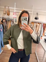 Chaqueta corta de textura efecto ante en color verde oliva, con diseño abierto, cuello redondo y bolsillos frontales amplios. Presenta mangas tres cuartos con puños vueltos y costuras decorativas horizontales.