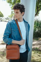 Bandolera de cuero color habano con textura granulada, cierre superior y bolsillo frontal abierto. Cuenta con una correa de tela ajustable con detalle de marca grabado.