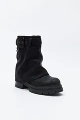 Bota de caña media color negro, con suela robusta dentada y detalle de hebilla ajustable en la parte superior. Presenta una argolla metálica lateral y un diseño de estética urbana con acabado texturizado.