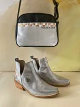 Bandolera de cuero negro y plateado con arte pintado a mano, perteneciente a la línea Ottenta de Carolina De Cunto. Cuenta con bolsillo frontal exterior, cierre superior con cremallera, forro interior con bolsillo y correa regulable con el logo de la marca.