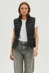 Chaleco acolchado negro, sin mangas, con cuello alto y cierre frontal con cremallera dorada. Presenta cordones ajustables a la altura de la cintura.