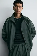 Chaqueta técnica de corte relajado en color verde oscuro, con cuello subido, cierre frontal de cremallera y detalles de líneas en contraste. Presenta bolsillos frontales con cremallera oculta y acabados elásticos en puños y ruedo.