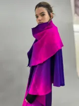 Prenda de abrigo tipo capa o chal de lana merino afieltrada, con diseño desestructurado y efecto degradé en tonos violeta y fucsia. Se ajusta al cuerpo mediante un nudo frontal.