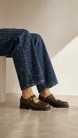 Mocasín de cuero color marrón con acabado texturizado, diseño de punta redonda con costuras artesanales visibles y cierre mediante una tira con hebilla lateral.