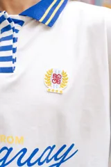Remera tipo polo de manga larga con diseño de mangas raglán a rayas horizontales azules y blancas. El cuerpo de la prenda es blanco, con cuello polo y una pequeña estampa en el pecho. Confeccionada en tejido de algodón con spandex.