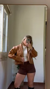 Campera corta de ecocuero color camel, con diseño de cuello solapa y cierre frontal con botones metálicos a presión. Presenta detalles de tachas metálicas a lo largo de la solapa y en el ruedo.