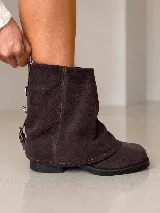 Bota corta de cuero gamuzado color marrón oscuro, con diseño de polaina superpuesta y detalle de hebillas metálicas en la parte posterior del talón. Cuenta con suela plana de goma y taco bajo.