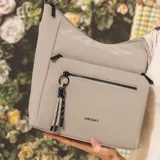 Cartera bandolera gris de formato amplio, con correa larga regulable y desmontable con inscripciones. Cuenta con cierre superior, bolsillo frontal con cierre y detalle colgante de cordones, y un bolsillo trasero con cierre.