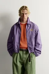 Cazadora regular fit de algodón color violeta con efecto lavado, cuello solapa y cierre de cremallera de doble dirección. Posee bordado frontal de gráficos Fruit of the Loom, bolsillos de vivo en la cadera y acabados con elástico.