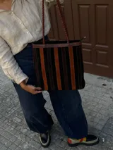 Cartera tipo tote de cuero con diseño de franjas verticales multicolores en tonos tierra, rojo y azul. Cuenta con correas trenzadas de cuero y cierre superior.