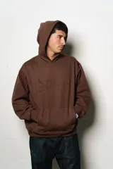 Hoodie oversize marrón oscuro con capucha y bolsillo canguro frontal. Presenta un logo bordado en tono gris claro en el pecho.