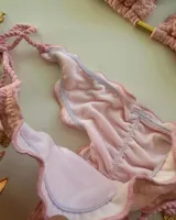 Bikini de dos piezas color rosa pálido con textura de pequeños puntos. El corpiño es triangular con bordes festoneados y un detalle de fruncido en el centro, sostenido por tirantes finos trenzados. La bombacha es de corte bajo con bordes festoneados y tiras laterales ajustables terminadas en pequeños dijes dorados.