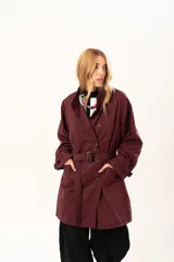 Trench de corte holgado color bordó, con cuello camisero, cierre frontal con botones y cinturón ajustable con hebilla. Presenta mangas largas con detalles de correas en los puños y bolsillos laterales.