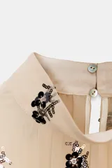 Blusa de cuello subido y manga larga con puños acampanados elásticos. Confeccionada en tejido semitransparente color beige con bordado de lentejuelas oscuras. Presenta cierre en la espalda con abertura y botones ocultos.