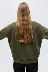 Buzo oversized color verde militar con interior cepillado suave. Presenta hombros caídos y un diseño de texto bordado en el pecho que dice "THE LAKE DISTRICT" con un pato. Tiene ribete acanalado en cuello, puños y bajo.