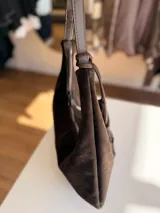 Cartera tipo hobo de textura gamuzada en color marrón oscuro, con diseño flexible y asa larga regulable. Presenta un detalle de flecos largos desmontables en uno de los laterales y cierre superior.