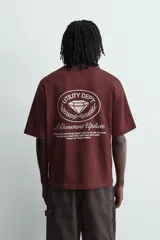 Camiseta de manga corta y cuello redondo, color granate oscuro, con estampado frontal en blanco que incluye texto y un escudo circular.