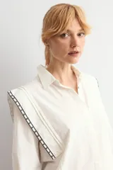 Camisa de color blanco con cuello clásico, cierre frontal de botones y mangas largas con puños. Presenta detalles de bordado geométrico en contraste a lo largo de los hombros y la parte superior de la espalda.