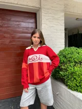 Sweater de punto rojo con rayas horizontales naranjas y blancas, cuello polo blanco y mangas largas. Presenta un diseño con letras curvas en el pecho.