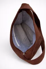 Bolso tipo bandolera de tamaño mediano, confeccionado en material con acabado efecto gamuza color chocolate. Presenta un diseño minimalista con cierre superior de cremallera y correa larga regulable con herrajes metálicos.
