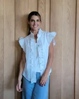 Musculosa blanca de tela bordada con detalles de volados en los hombros y a lo largo de la botonadura frontal. Presenta cuello alto tipo mao y cierre con botones nacarados.