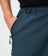 Pantalón de corte holgado en color azul petróleo, confeccionado en microfibra de poliéster. Presenta un diseño utilitario con múltiples bolsillos funcionales, cierres visibles y detalles técnicos en las piernas.