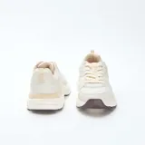 Zapatillas estilo running de color beige, confeccionadas con una combinación de paneles de cuero sintético y malla transpirable. Presentan cordones al tono, suela robusta con relieve y detalles en color marrón oscuro en la base.