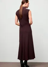 Vestido largo de corte evasé en color marrón oscuro, con cuello alto y cierre de botones en la parte posterior. Presenta un diseño sin mangas con tirantes anchos y una costura diagonal en la cintura que aporta estructura a la prenda.