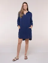 Vestido corto blanco de algodón, marca Dash, con cuello camisero, escote en V y bolsillos laterales. Presenta mangas tres cuartos y calce holgado con ruedo irregular.
