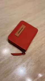 Billetera de cuero color rojo con cierre perimetral y etiqueta frontal de cuero en tono natural con el logo de la marca grabado.
