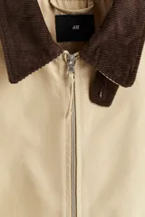 Campera tipo blouson de algodón color beige claro, con cuello de pana marrón oscuro. Posee cierre frontal, bolsillos delanteros ribeteados y elástico ajustable con cordón en el bajo.