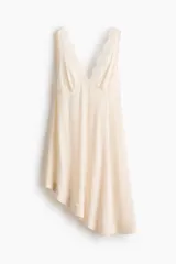 Vestido midi color beige de viscosa, sin mangas, con escote en V profundo adelante y atrás decorado con ribetes de encaje. Presenta una falda acampanada con bajo asimétrico y costuras en zigzag.