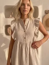Vestido corto de lino color beige, sin mangas con volados en los hombros, cuello mao con botones frontales de nácar, corte en la cintura fruncido y falda con volado en el ruedo.