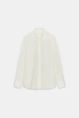 Camisa de popelín color crudo con cuello solapa y aplicación desmontable de pelo sintético en tono marrón. Presenta cierre frontal con botones, manga larga con puños y bajo asimétrico.