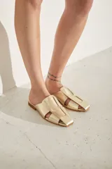 Zueco plano de cuero color dorado, con diseño destalonado, puntera cuadrada y tiras anchas entrelazadas en el empeine.