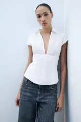 Top de popelina blanca de corte corsetero, con escote en pico pronunciado, cuello tipo solapa, mangas muy cortas y costuras marcadas.