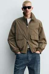 Cazadora regular fit de tejido técnico color marrón, con cuello solapa, manga larga y cierre frontal de cremallera. Presenta bolsillos de plastrón con solapa en la cadera y acabados con rib elástico.
