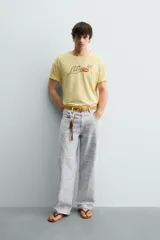 Camiseta de corte relajado color amarillo con efecto lavado. Presenta cuello redondo, mangas cortas con dobladillo y un estampado frontal con texto y un parche decorativo.