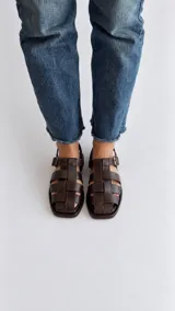 Sandalias de cuero color marrón, estilo romano con tiras entrelazadas, punta cuadrada y cierre de hebilla metálica en el tobillo.