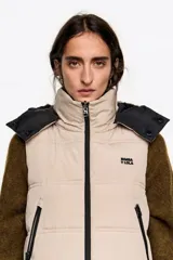 Chaleco acolchado de largo medio y corte en línea A, confeccionado en tejido técnico color beige con detalles en negro. Presenta cuello subido, capucha desmontable, cierre frontal de cremallera, bolsillos laterales con cremallera, logo bordado en el pecho y aberturas laterales con cremallera.