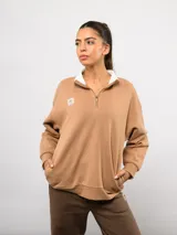 Buzo de felpa color tostado con cuello alto y medio cierre frontal. Presenta un diseño de corte holgado con hombros caídos, puños y ruedo acanalados, y el logo de la marca bordado en el pecho.